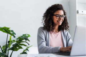 mulher negra de óculos sorrindo enquanto está sentada a uma mesa usando um computador
