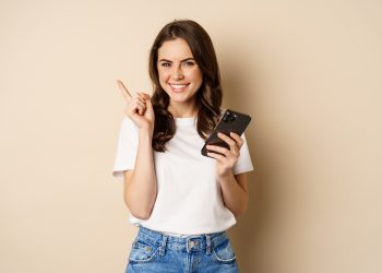 elegante jovem moderna segurando o celular apontando o dedo para a esquerda e sorrindo o conceito de compras scaled