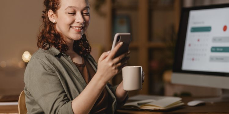 woman home using smartphone front computer while having coffee scaled