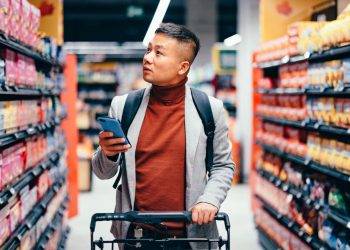 Um homem no supermercado com um carrinho e o celular na mão