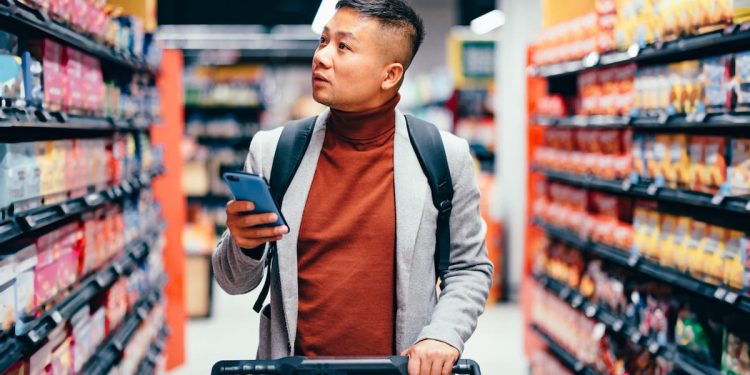 Um homem no supermercado com um carrinho e o celular na mão