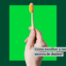 Mão de pessoa segurando uma escova de dente com cerdas laranjas em um fundo verde escrito como escolher a melhor escova de dentes?