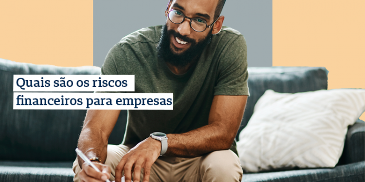 Homem sorrindo, sentado em um sofá, inclinado para frente escrevendo em um papel apoiado na mesa de centro. Na imagem há o texto "quais são os riscos financeiros para empresas".