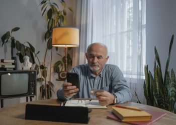 Idoso segurando carteirinha de plano e celular sentado em mesa de sala.