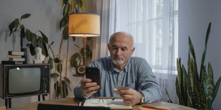 Idoso segurando carteirinha de plano e celular sentado em mesa de sala.