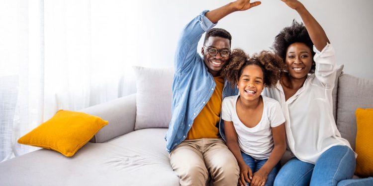 Família feliz em casa no sofá fazendo sinal de casa com as mãos.