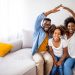 Família feliz em casa no sofá fazendo sinal de casa com as mãos.