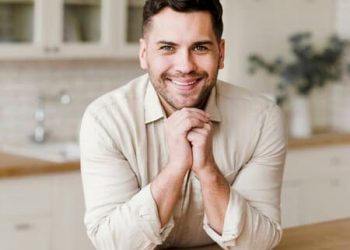 Um homem na sua cozinha, apoiando em seu balcão, levando suas duas mãos ao queixo.