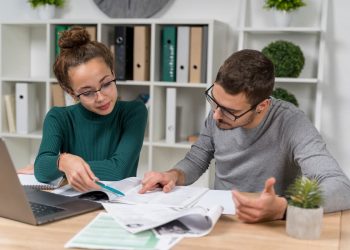 Um casal analisando uns papéis com um computador em cima da mesa