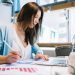 Uma mulher em sua mesa, analisando as informações que estão em seu computador e escrevendo no papel