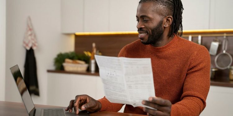 Um homem segurando em sua mão um papel e mexendo ao mesmo tempo em seu computador