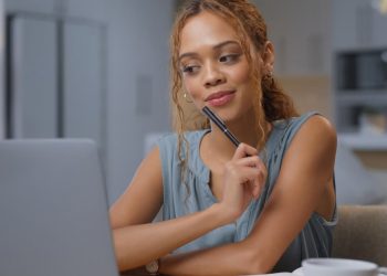 Uma mulher de regata azul, sentada em seu escritório, olhando para seu computador e com o lápis apoiado em seu queixo