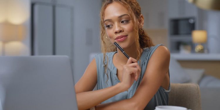 Uma mulher de regata azul, sentada em seu escritório, olhando para seu computador e com o lápis apoiado em seu queixo