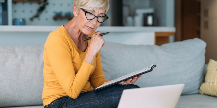 Uma senhora de blusa amarela e cabelo branco, sentada em um sófa, com papéis e caneta em suas mãos, e olhando para o computador que está na mesa na sua frente