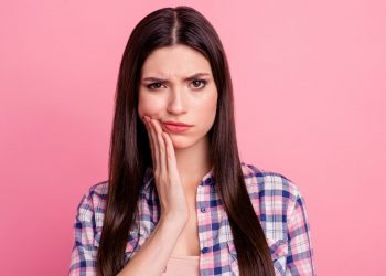 Uma mulher de camisa quadriculada em um fundo rosa, com a mão em sua bochecha com o rostro mostrando incomodo