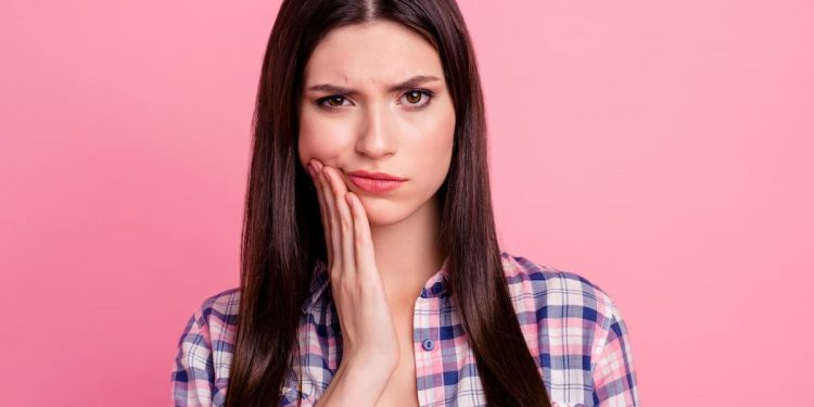 Uma mulher de camisa quadriculada em um fundo rosa, com a mão em sua bochecha com o rostro mostrando incomodo