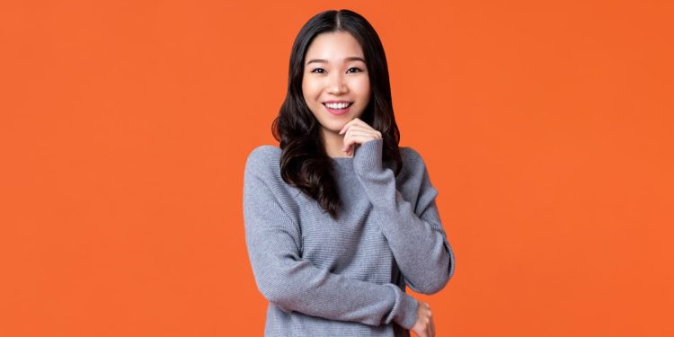 Uma mulher com blusa cinza, com a mão em seu queixo, rindo para a foto