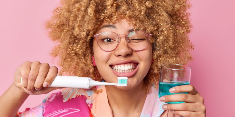 mulher de cabelos cacheados sorri com escova de dente na frente da boca e enxaguante bucal na outra mao