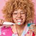 mulher de cabelos cacheados sorri com escova de dente na frente da boca e enxaguante bucal na outra mao