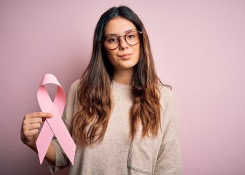 mulher de cabelos longos e óculos de grau, segurando fita rosa que representa o outubro rosa