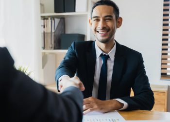Homem em escritório de terno dando as mãos com contrato.