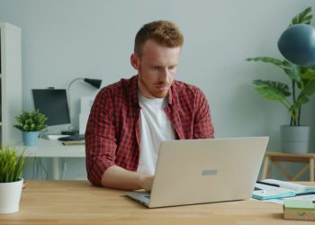 Homem sentado em mesa de escritório mexendo em notebook.