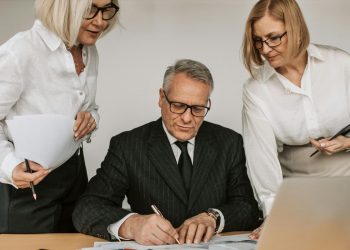 Homem mais velho assinando documento em mesa com duas mulheres de meia idade auxiliando ao lado.