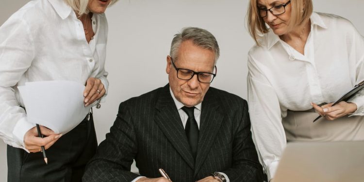Homem mais velho assinando documento em mesa com duas mulheres de meia idade auxiliando ao lado.