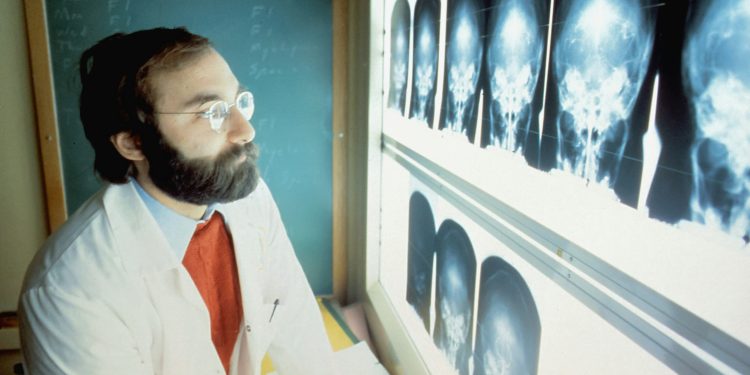 Homem de óculos vestido com avental olhando para radiografias odontológicas.