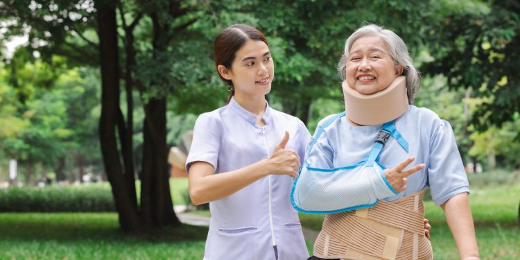 Profissional da saúde e paciente com braço enfaixado caminhando em área verde, com árvores ao fundo.