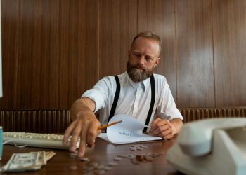Homem loiro de barba fazendo contas financeiras sentado em mesa marrom.