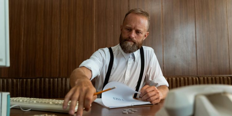 Homem loiro de barba fazendo contas financeiras sentado em mesa marrom.