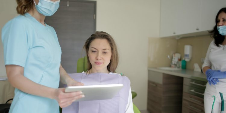 Doutora mostrando tablet para paciente sentada dentro da sala de atendimento odontológico.