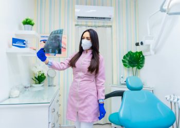 Mulher olhando para radiografia dental dentro de sala branca, vestida de rosa com máscara.