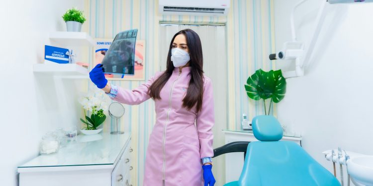 Mulher olhando para radiografia dental dentro de sala branca, vestida de rosa com máscara.